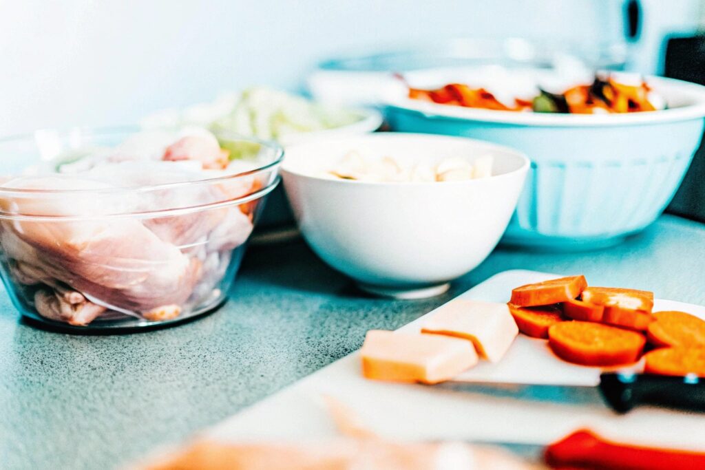 A kitchen counter with raw chicken breasts, bowls of fresh vegetables, and a cutting board with sweet potatoes being diced