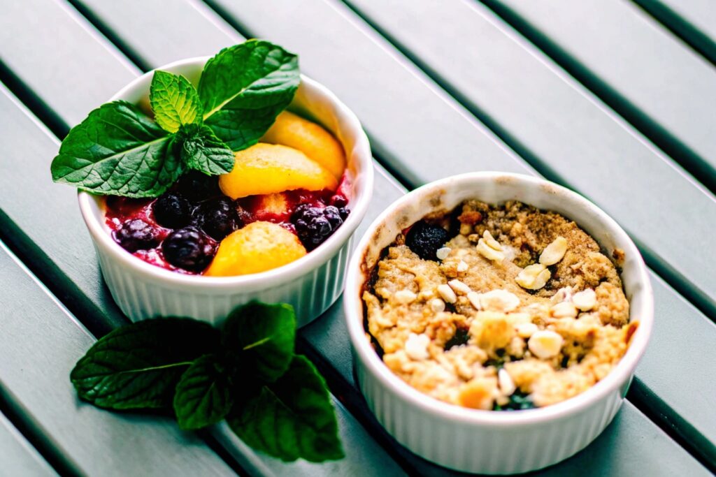 Two ramekins side by side; one with peach cobbler showcasing a biscuit topping, and one with peach crumble featuring a crunchy streusel topping