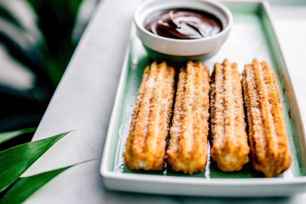 A plate of golden churros dusted with cinnamon sugar, accompanied by a small bowl of thick chocolate sauce