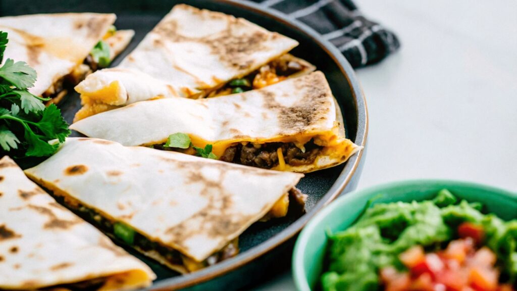 Thinly sliced, juicy steak arranged on a wooden cutting board, surrounded by fresh tortillas, shredded cheese, and a bowl of salsa