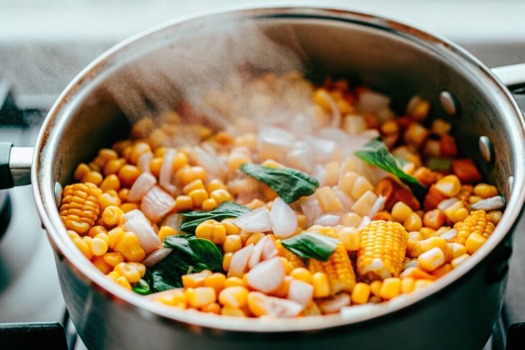 A pot of fresh corn kernels simmering with onion and garlic on a stovetop, steam rising