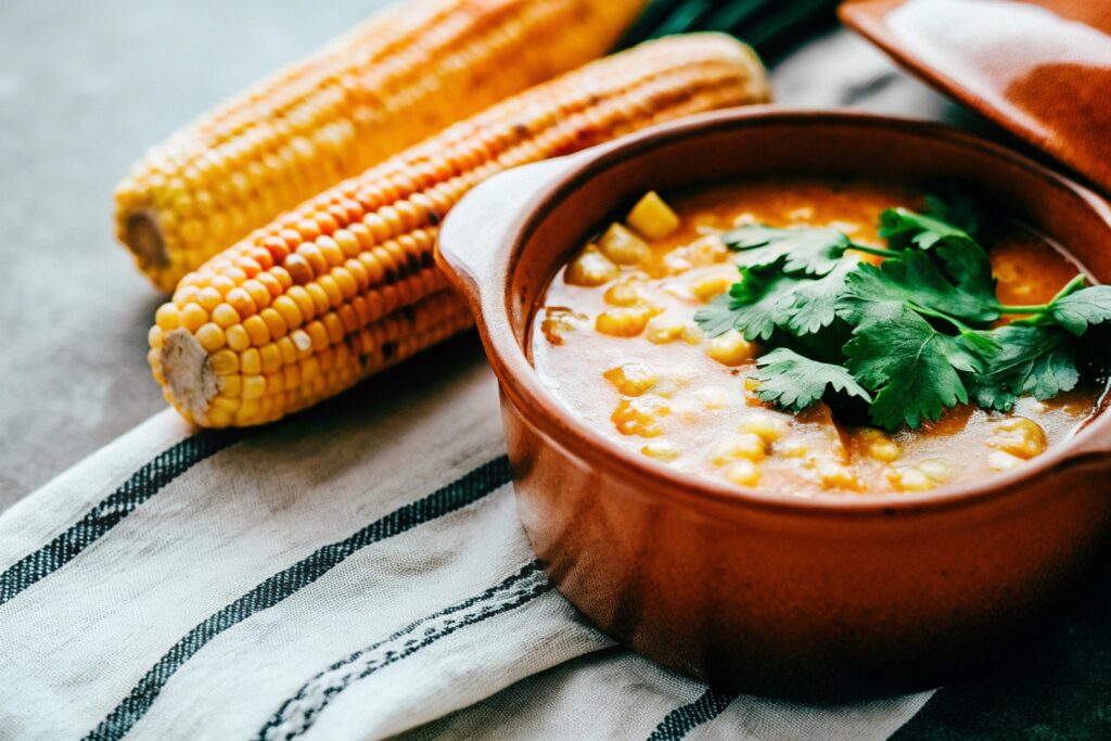 A rustic kitchen scene featuring a pot of corn soup surrounded by fresh corn cobs, cilantro, and traditional Salvadoran textiles