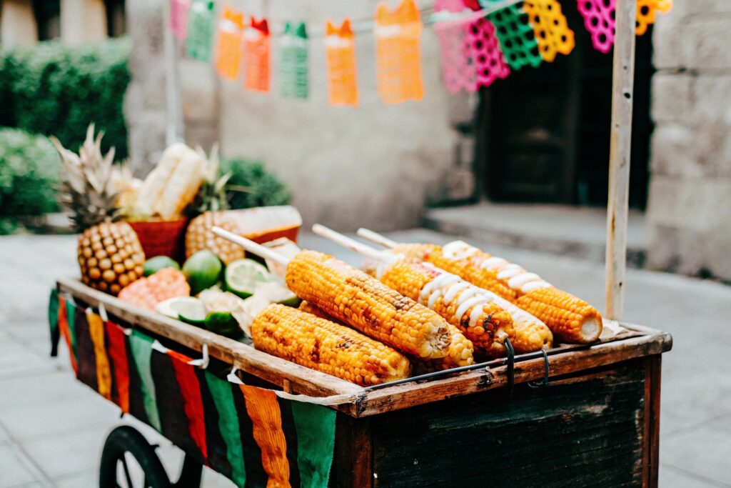 A rustic street cart in Mexico selling grilled elote, surrounded by vibrant produce and traditional Mexican decorations.