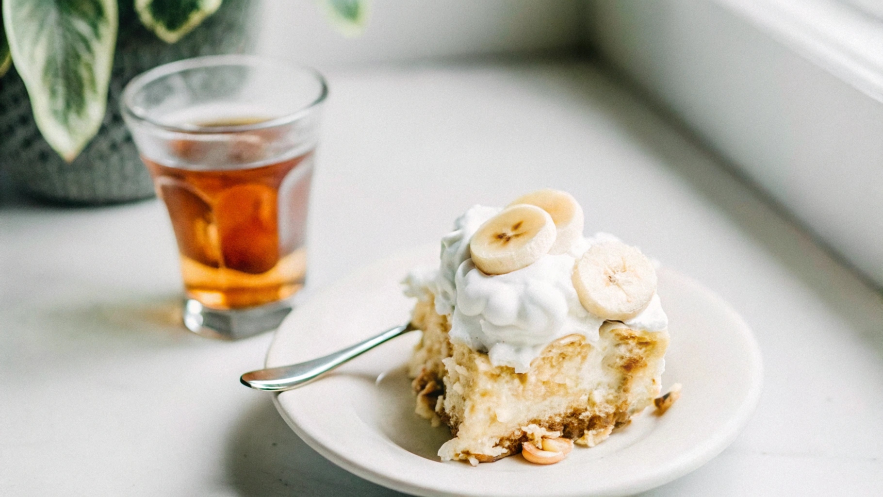 A slice of banana pudding on a white plate with a spoon, accompanied by a glass of iced tea on a wooden table, evoking classic Southern charm