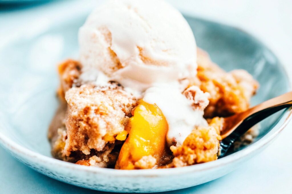 A serving of peach cobbler with cake mix topping on a white plate, garnished with a scoop of melting vanilla ice cream