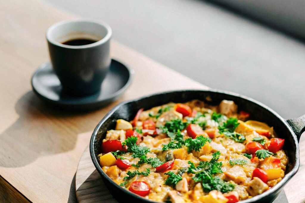 A skillet of chicken and egg scramble with parsley garnish, set on a wooden table with a cup of coffee.