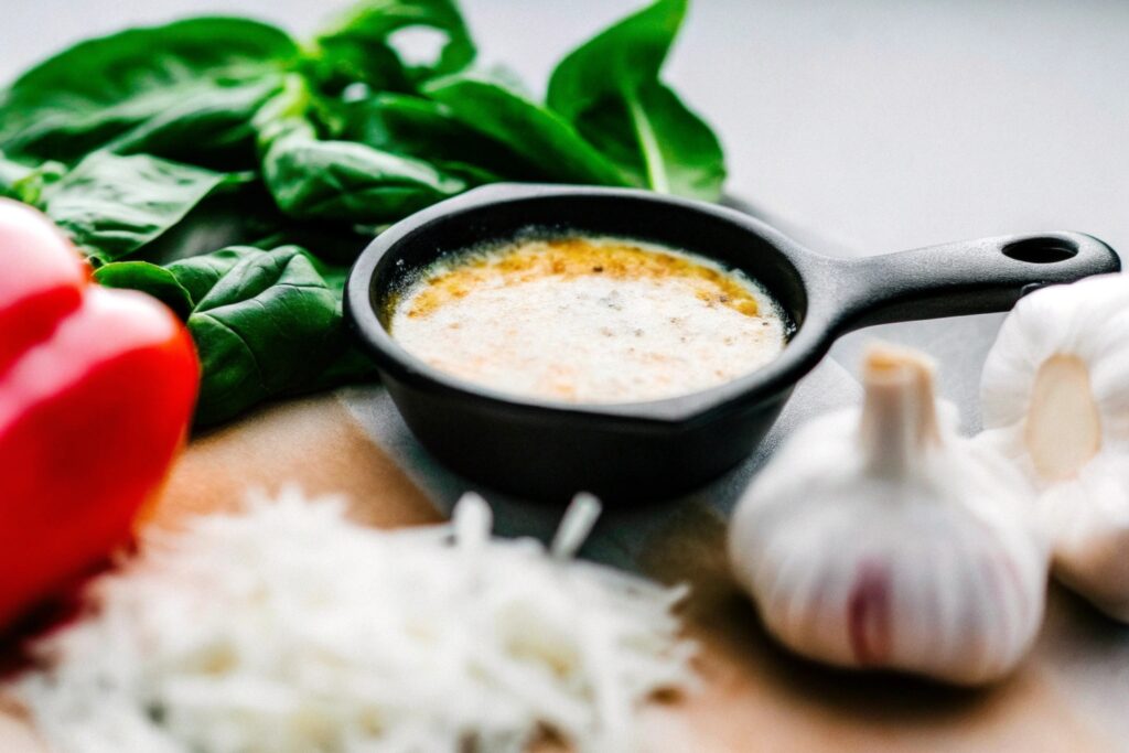 A small skillet containing leftover Alfredo sauce, surrounded by fresh ingredients like grated Parmesan cheese, a clove of garlic, and a sprig of parsley