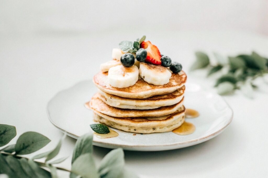 A stack of golden banana oat pancakes topped with fresh banana slices, mixed berries, and a drizzle of honey