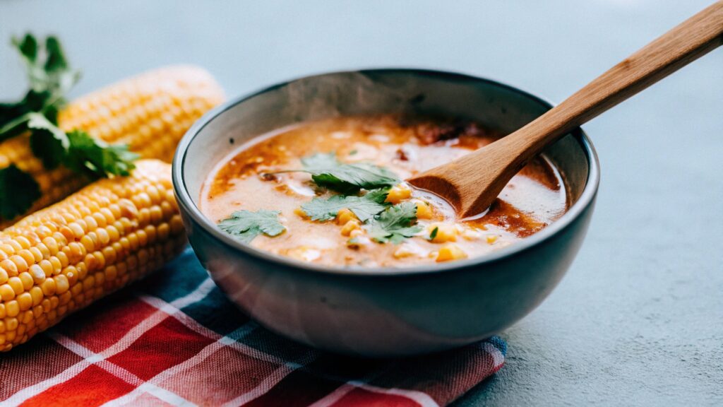 A steaming bowl of El Salvadoran corn soup with a wooden spoon, surrounded by fresh corn cobs, cilantro, and a colorful Salvadoran tablecloth