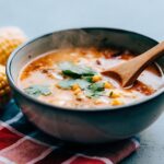 A steaming bowl of El Salvadoran corn soup with a wooden spoon, surrounded by fresh corn cobs, cilantro, and a colorful Salvadoran tablecloth