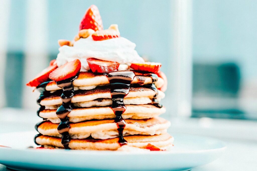 A tall stack of chocolate pancakes drizzled with syrup and garnished with strawberries and whipped cream