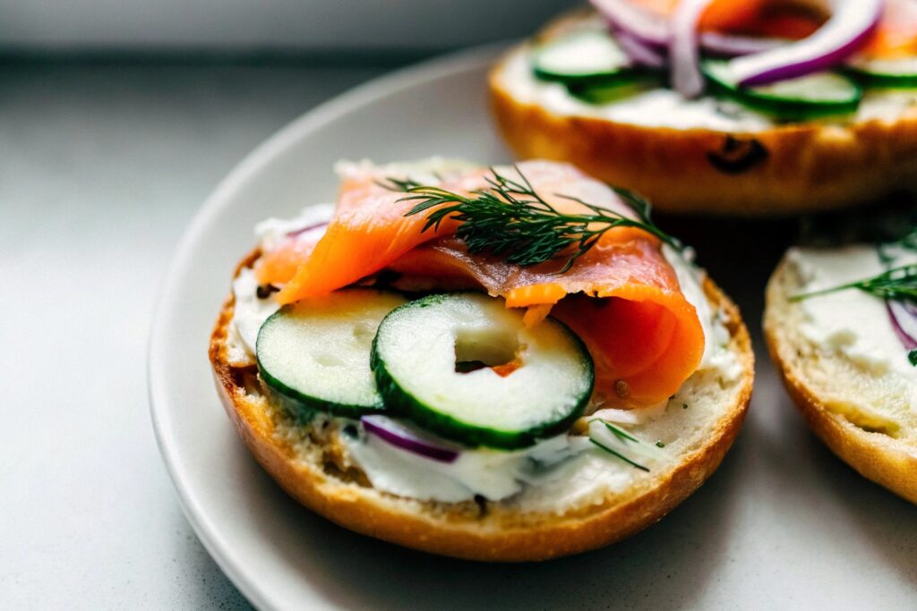 A toasted bagel topped with creamy cheese, smoked salmon slices, red onion rings, cucumber slices, and garnished with fresh dill, presented on a white plate