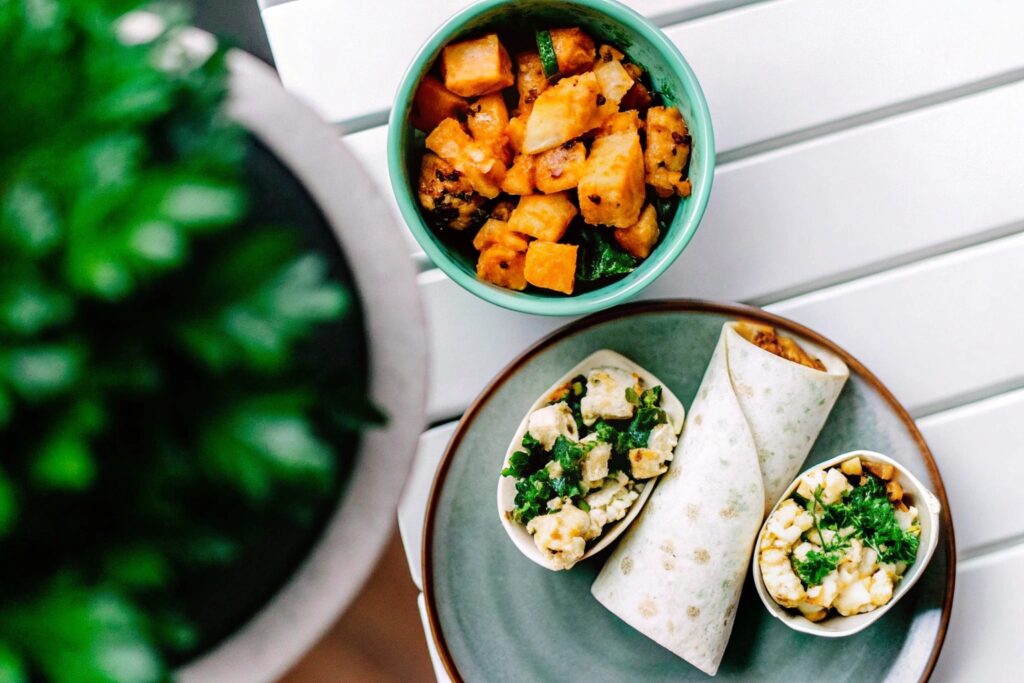 A top-down view of a wooden table with a plated chicken breakfast scramble, a vibrant sweet potato bowl, and a wrapped chicken tortilla, ready to serve