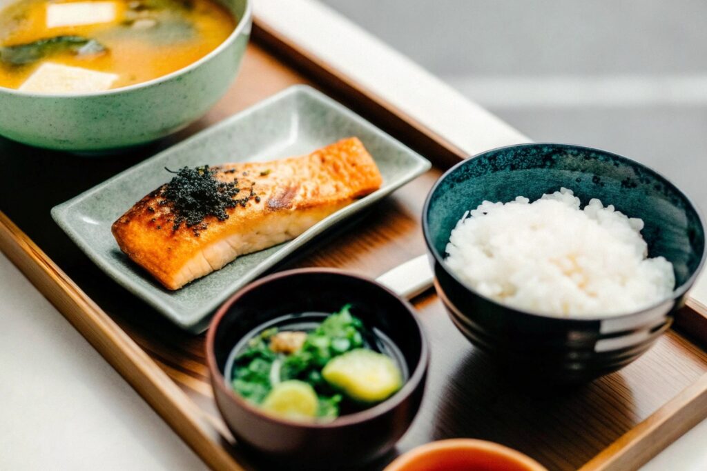 A traditional Japanese breakfast spread featuring grilled salmon, steamed rice, miso soup, pickles, and nori on a wooden table