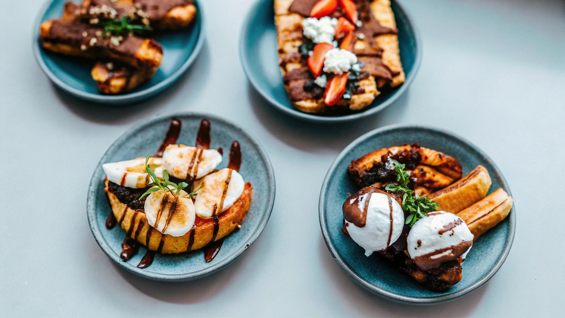 A variety of plated fried banana dishes, including a dessert version drizzled with chocolate, a breakfast option topped with yogurt and granola, and a savory side garnished with fresh herbs