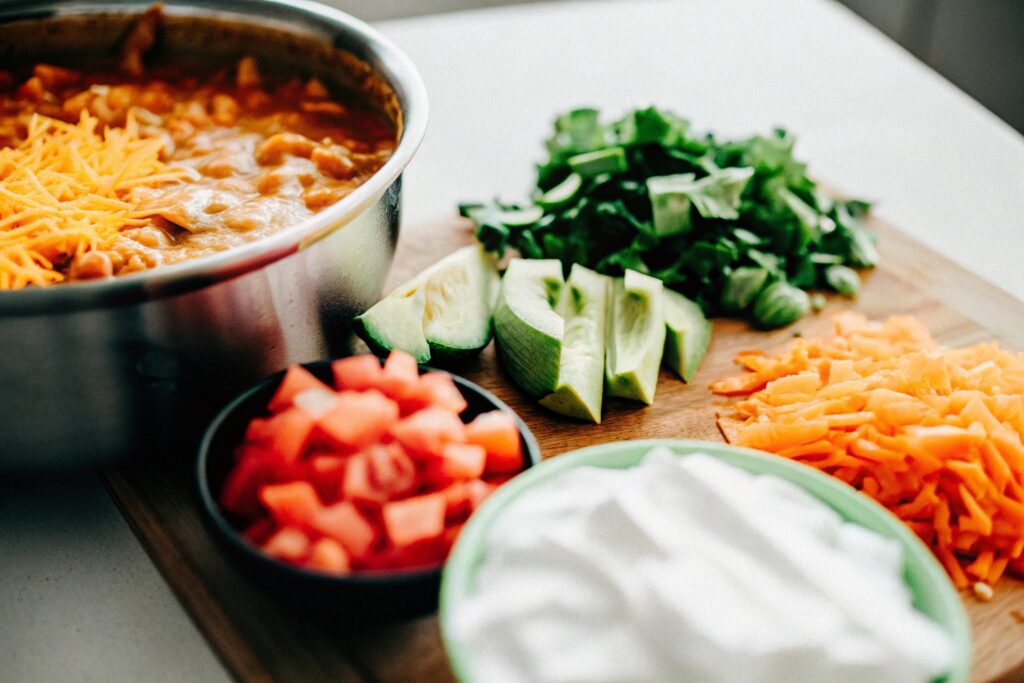 A variety of toppings and ingredients displayed alongside a pot of taco soup, including shredded cheese, avocado slices, crushed tortilla chips, and fresh cilantro