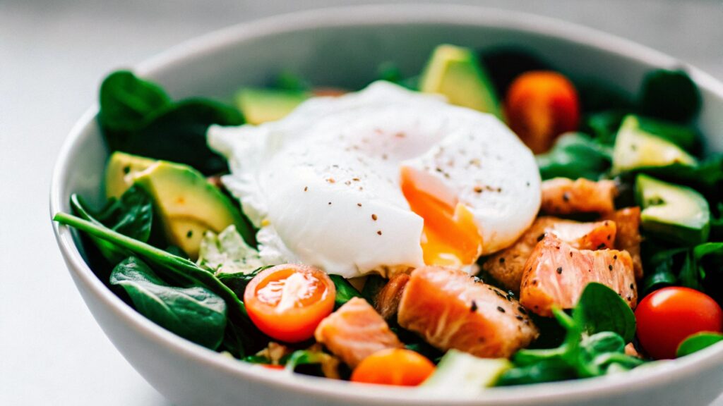 A vibrant breakfast bowl featuring a poached egg, grilled salmon, avocado, and cherry tomatoes on a bed of mixed greens.