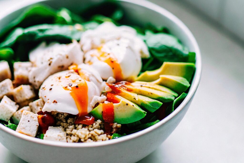 A breakfast bowl with diced chicken, quinoa, poached eggs, spinach, and avocado, topped with hot sauce drizzle.