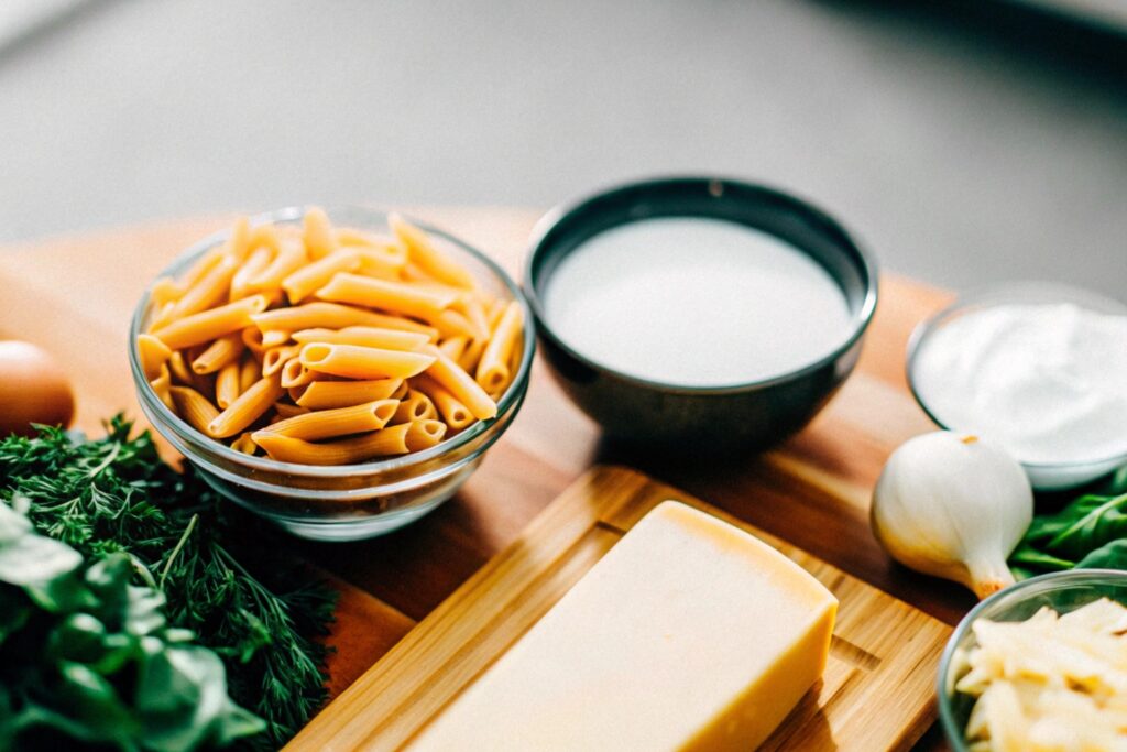 A vibrant display of ingredients for penne Alfredo, including uncooked penne pasta, a block of Parmesan cheese, butter, cream, garlic cloves, and fresh parsley on a rustic wooden counter.