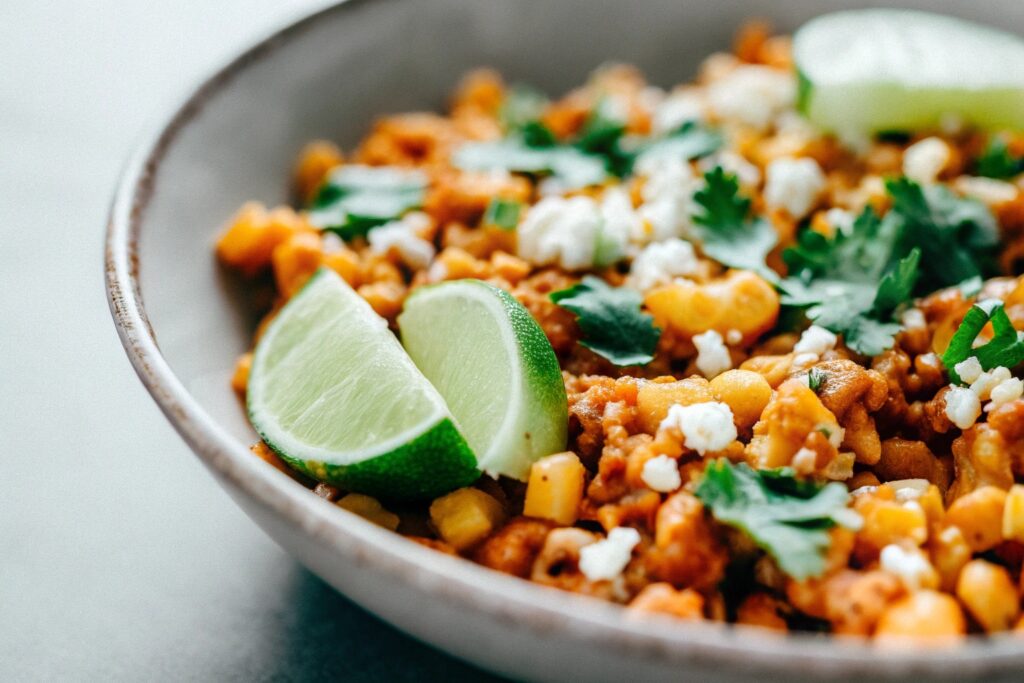 A vibrant plate of Mexican corn topped with ground chicken, garnished with fresh cilantro, lime wedges, and cotija cheese crumbles.