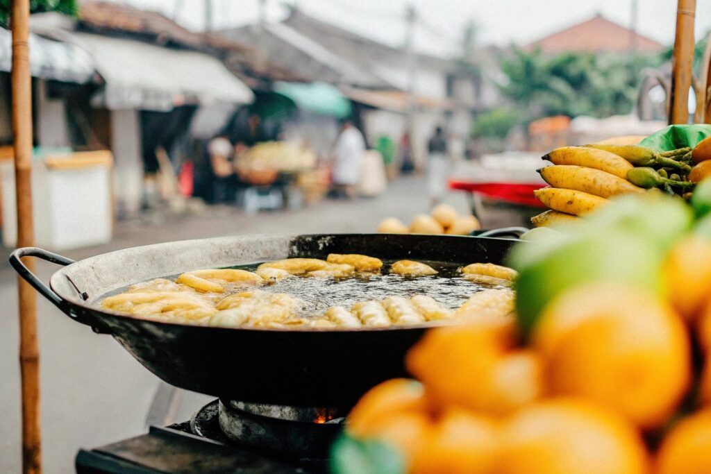 A vibrant street food stall in Southeast Asia showcasing fried bananas being prepared in a large wok, surrounded by fresh tropical fruits.