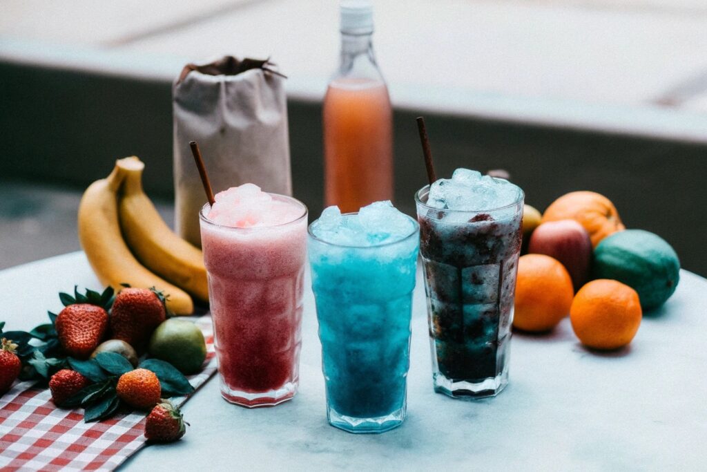 A vibrant table featuring colorful slushy drinks in clear glasses, surrounded by fresh fruits, syrups, and a bag of crushed ice