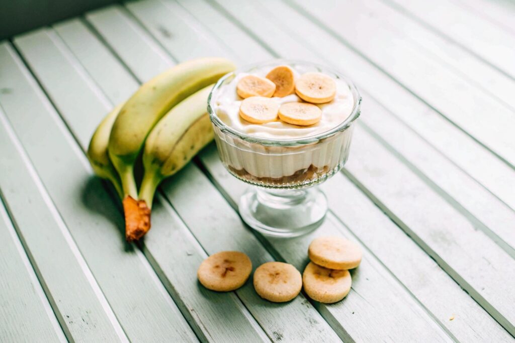 Vintage-style banana pudding served in a trifle dish, surrounded by fresh bananas and vanilla wafers on a rustic wooden table.