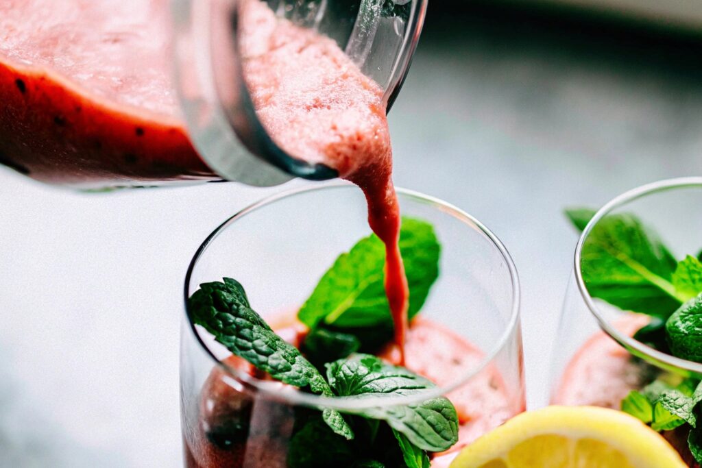 A close-up shot of a blender pouring a freshly made berry slushy into a glass, garnished with mint leaves and a slice of lemon