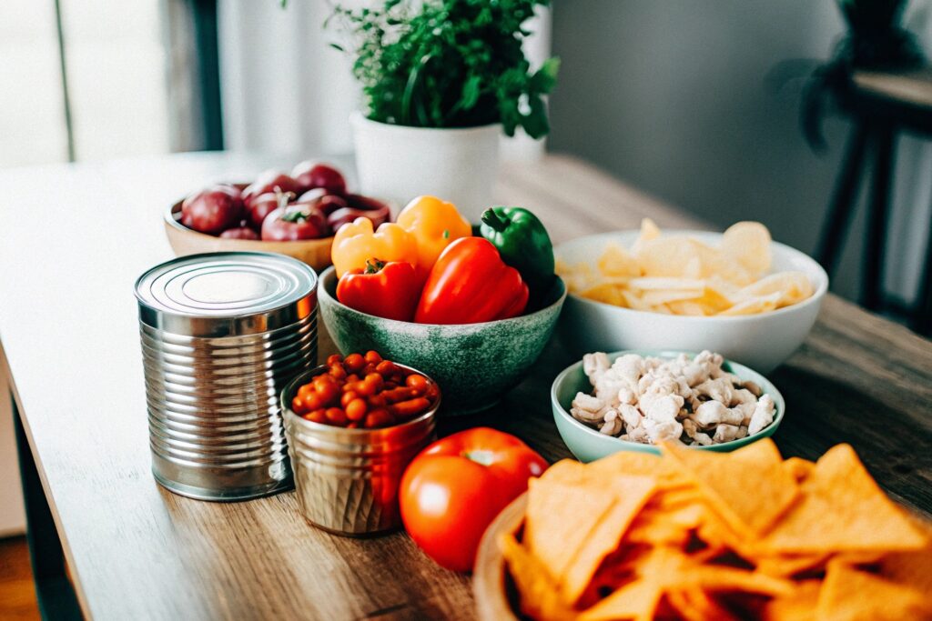 Ingredients for chicken taco soup arranged on a wooden table, including fresh vegetables, canned goods, taco seasoning, and raw chicken breasts.