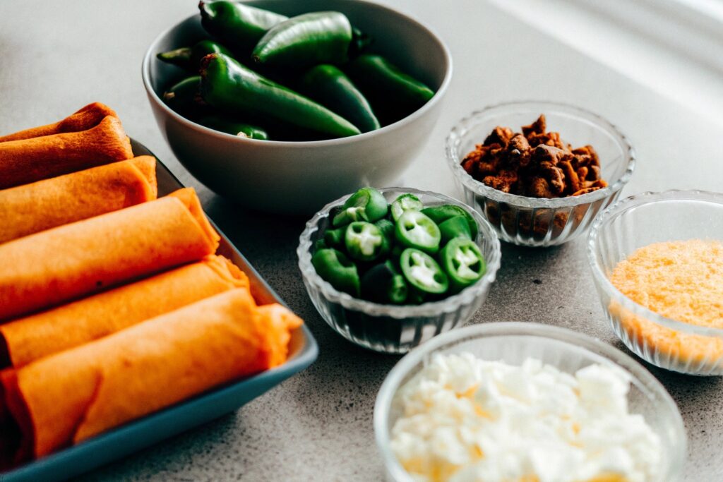 Ingredients neatly arranged on a countertop: egg roll wrappers, jalapeños, cream cheese, shredded cheese, and spices in small bowls