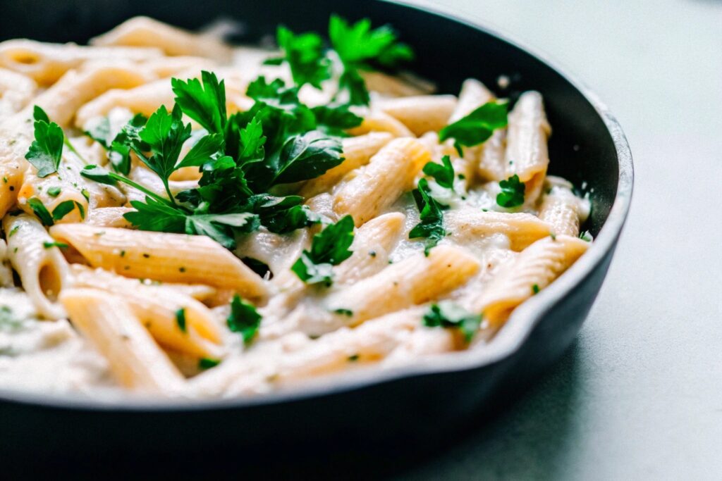 A skillet filled with penne pasta coated in creamy Alfredo sauce, garnished with freshly chopped parsley, placed on a wooden table.