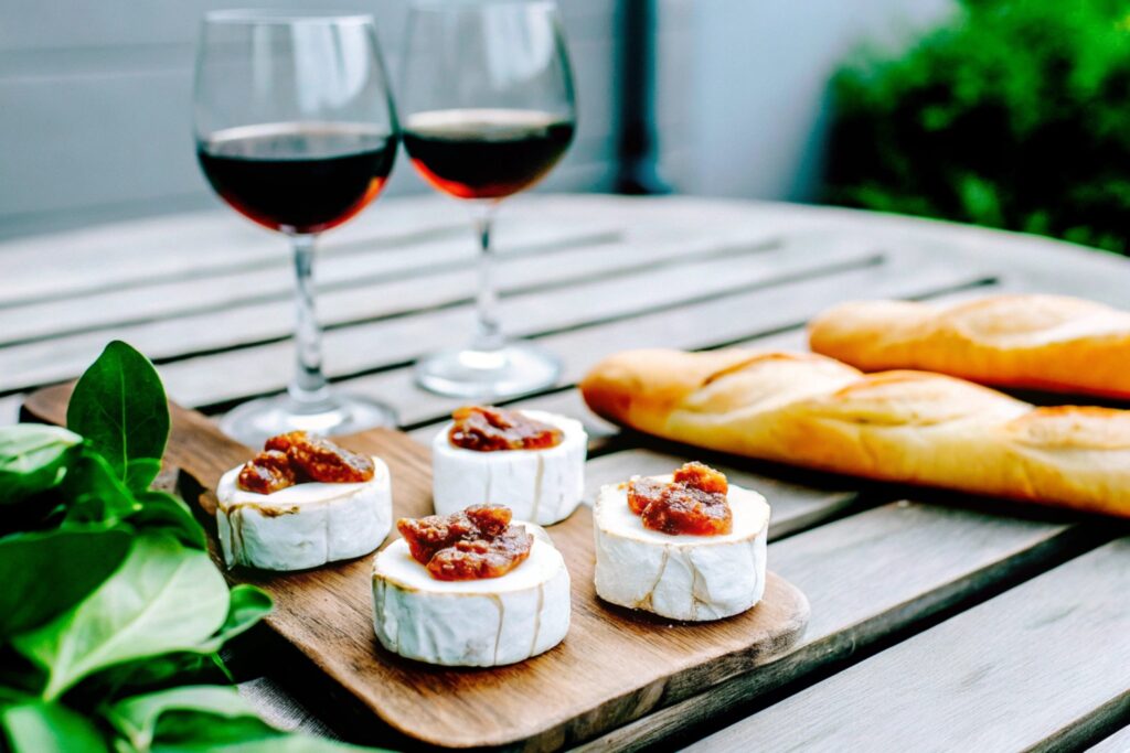 Brie bites topped with fig jam on a rustic wooden table, surrounded by glasses and slices of French baguette, highlighting their cultural roots.