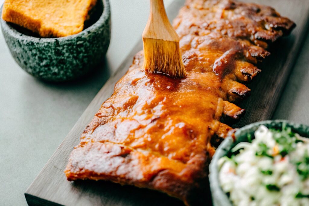 A slab of smoked ribs being brushed with BBQ sauce, surrounded by side dishes like cornbread, coleslaw, and pickles