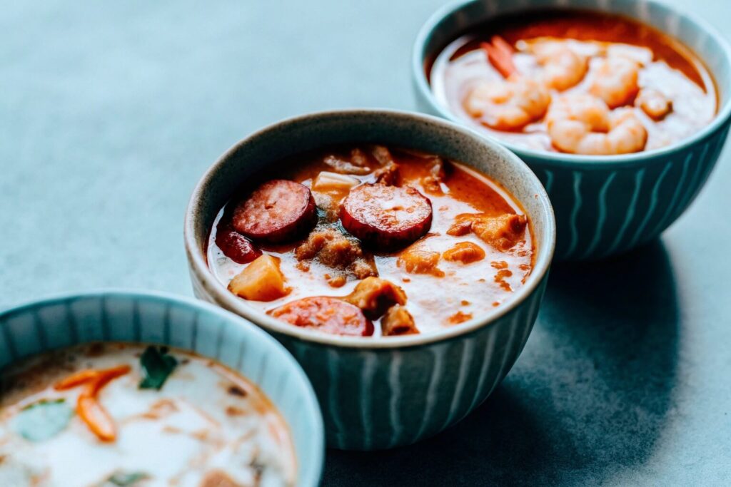 A close-up of different variations of swamp soup in bowls—one with shrimp, one with sausage, and one creamy version with a swirl of coconut milk